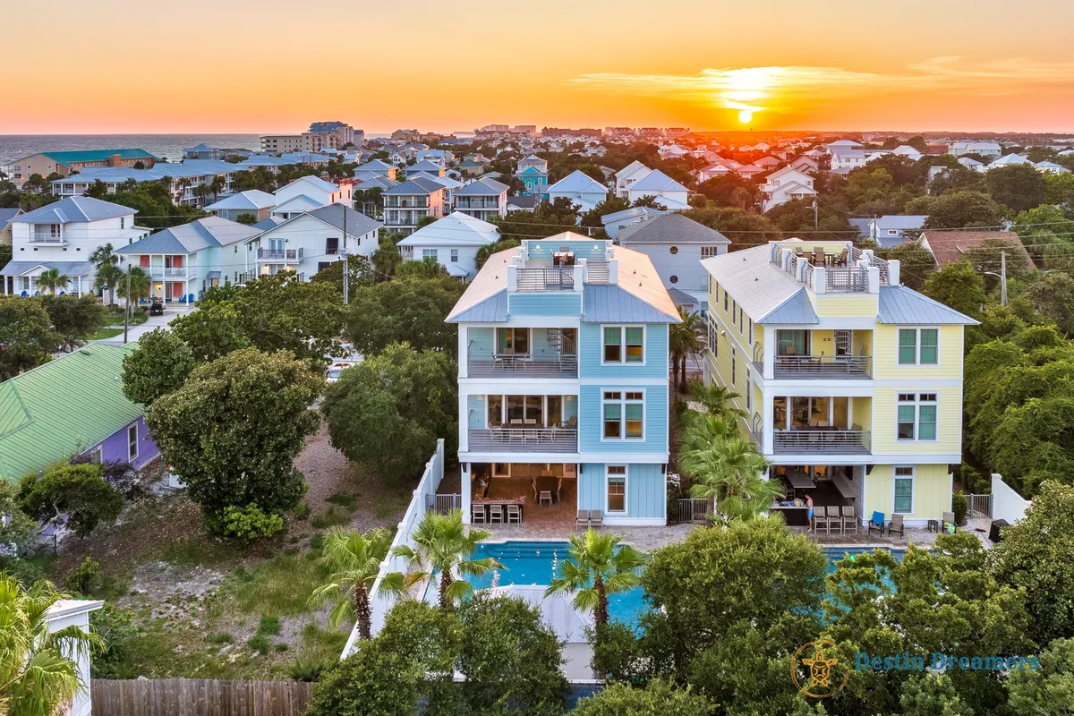 Two colorful modern homes flank a sparkling swimming pool, surrounded by lush greenery and a dramatic sunset sky. The scene radiates social energy, with chairs and a garden that invite gatherings and relaxation after a day of excursions.