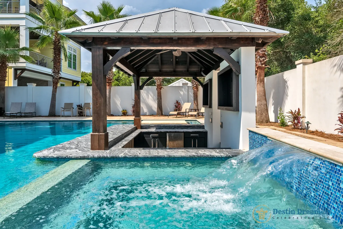 A vibrant exterior pool area featuring a beautiful gazebo, a shimmering pool, a waterfall feature, and lush palm trees. The space is set up for sunbathing, swimming, and entertaining, providing a picturesque setting for dogs and their owners to spend time outside together.