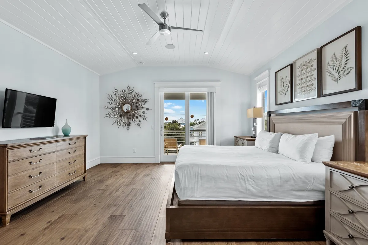 A serene king suite featuring a king-sized bed with crisp white linens, a wooden dresser, a large sunburst mirror, a wall-mounted television, and sliding glass doors that open onto a private balcony. Natural light fills the room, highlighting its spaciousness and inviting atmosphere.