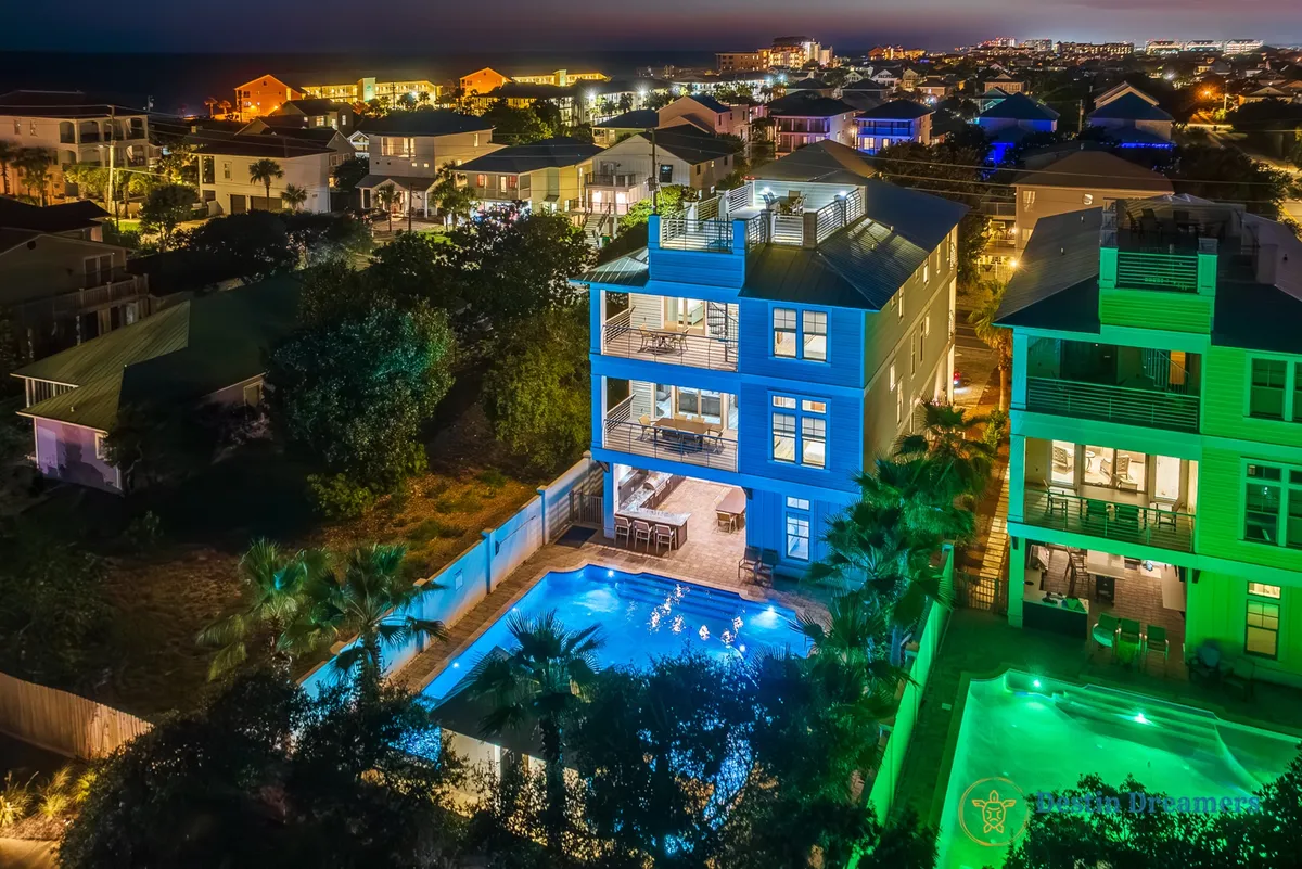 A striking exterior view of the home at night, with the modern architecture illuminated and the inviting swimming pool in the foreground. The scene captures guests enjoying the outdoor space, with the rooftop terrace visible above and the Gulf providing a dramatic sunset backdrop.