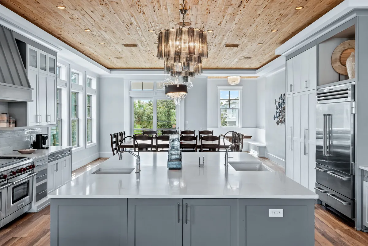 This image depicts a spacious, elegant kitchen and dining area featuring a large kitchen island, statement chandelier, and abundant natural light streaming through expansive windows. The dining area is designed for entertaining and group meals, with ample seating and a layout that invites social interaction and collaborative cooking.