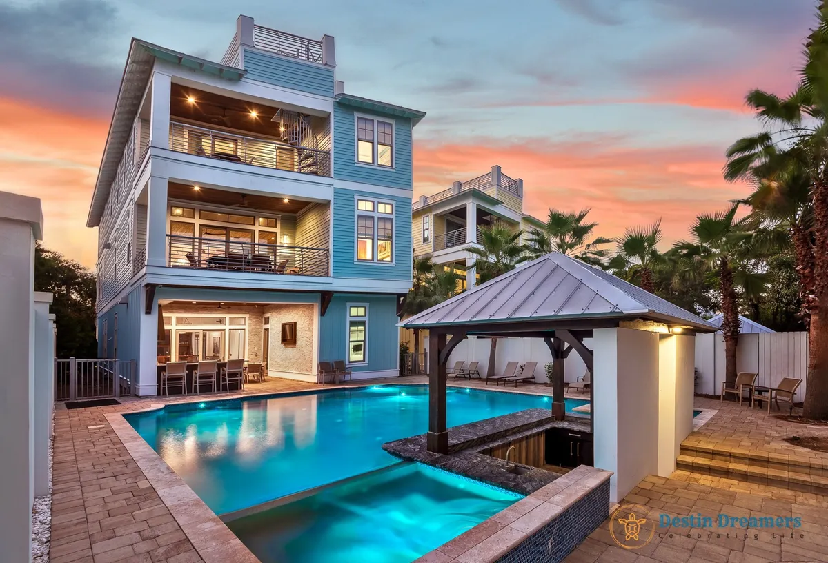 The image shows a stunning multi-story beach house in Destin, Florida, with spacious balconies overlooking a private heated pool featuring a swim-up bar and a poolside tiki kitchen. The scene is set at sunset, casting a warm and inviting glow over the property, highlighting spaces perfect for outdoor dining, lounging, and entertaining.