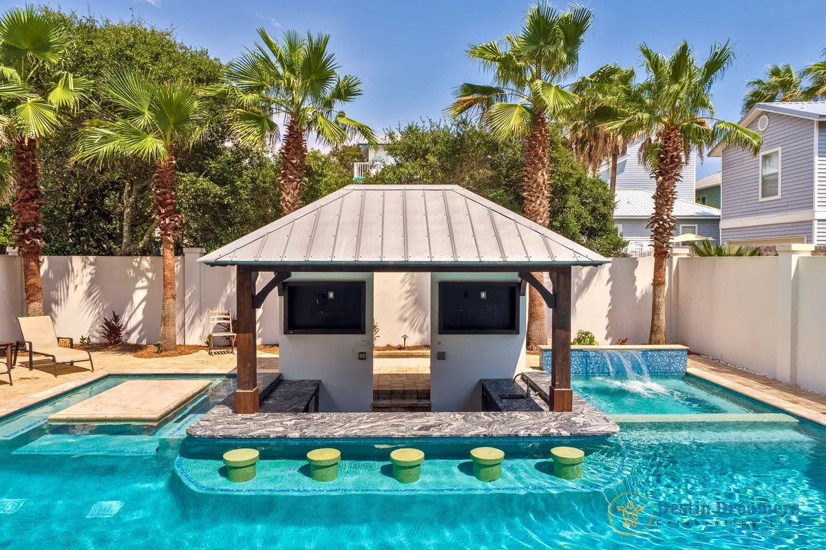 A vibrant exterior shot featuring a private heated pool with a sunken swim-up bar under a tiki-style pavilion, surrounded by lush palm trees and lounge chairs. The pool's waterfall feature adds a festive ambiance, and the area is well-lit for both daytime and evening celebrations.