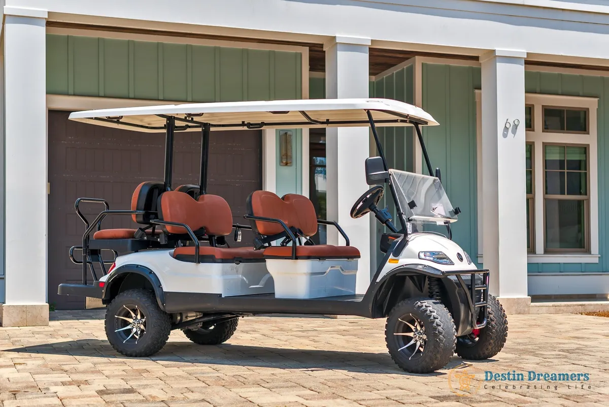 A 6-passenger street-legal golf cart is parked outside a modern beach home with white pillars and light green paneling. The paved driveway and visible entrance suggest easy access for outings. The golf cart is ready for family or group explorations, including trips to local parks, pet-friendly patios, and neighborhood adventures.