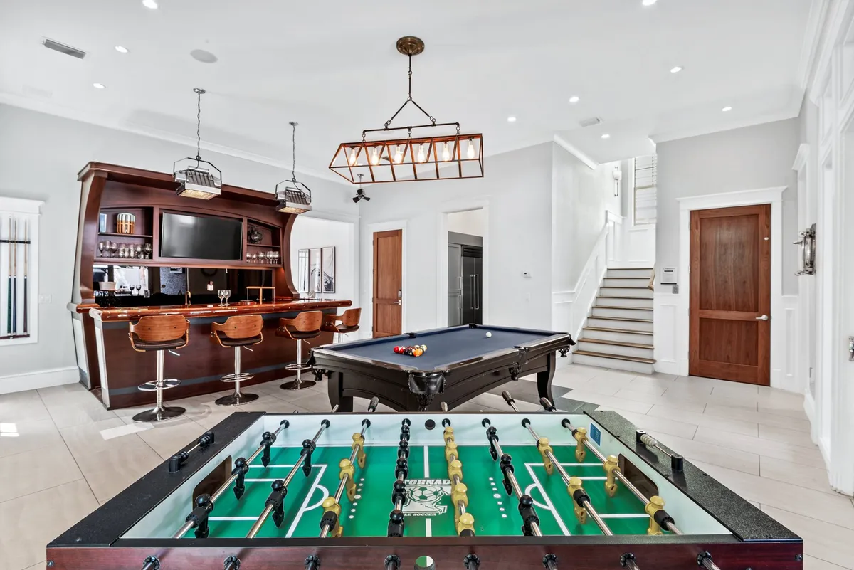An inviting indoor bar and game room with a pool table and foosball table, bar seating, a flatscreen TV, and a staircase leading to upper floors. The space is styled for lively social interaction, with warm lighting that encourages evening gatherings and friendly competition.