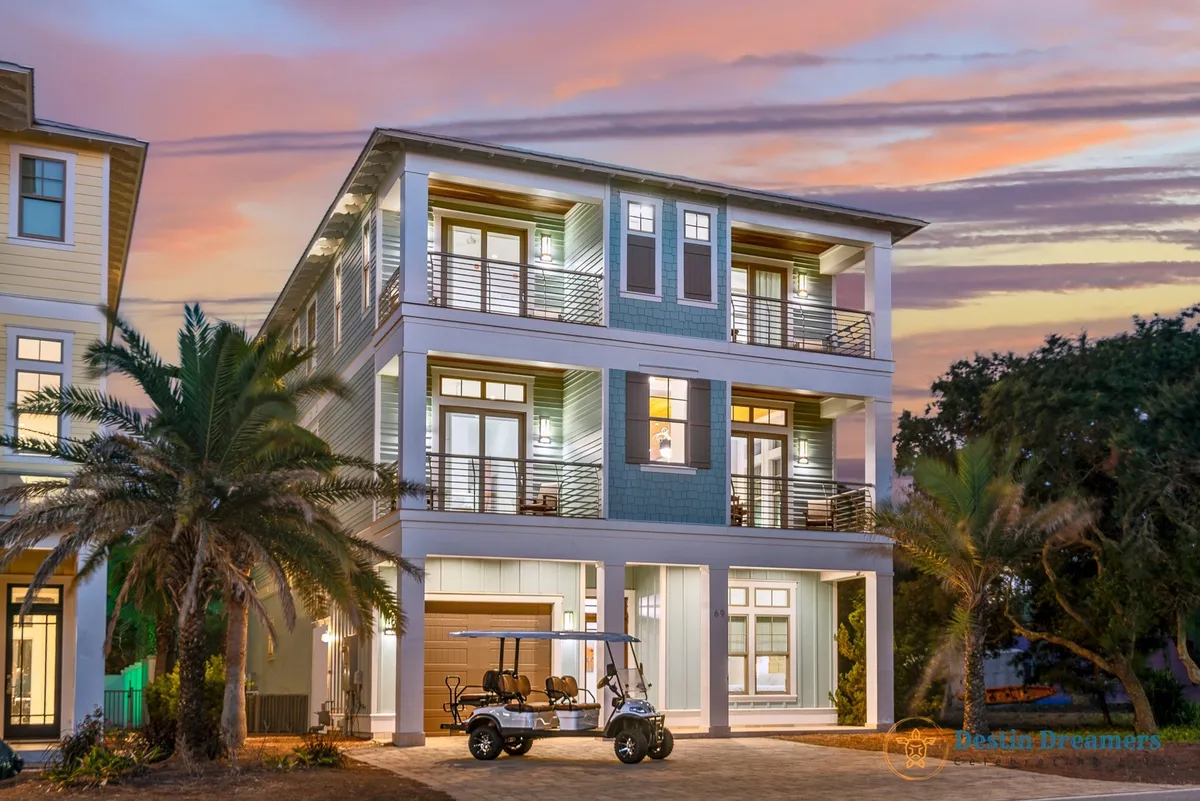 This image features a three-story modern beach house with multiple balconies, a 6-passenger street-legal golf cart parked in the driveway, and lush palm trees framing the property. The colorful sunset sky enhances the sense of arrival and adventure.