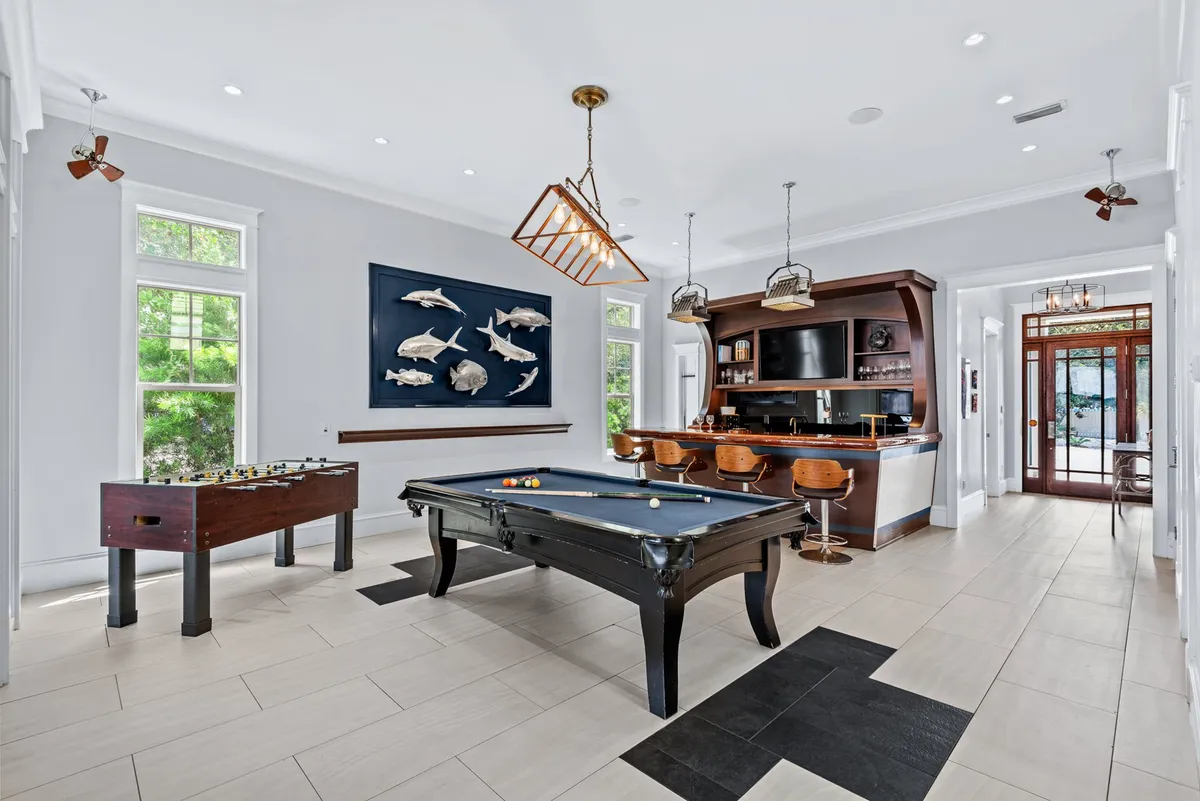 A social interior scene with a pool table, foosball table, a bar with TV and bar stools, and decorative fish wall art. The arrangement encourages friendly competition, casual drinks, and group interaction in a playful coastal setting.