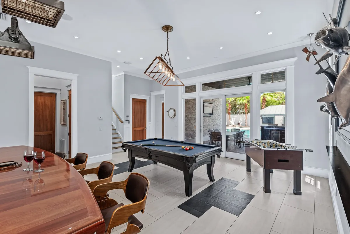 A vibrant game room featuring a centrally placed pool table, a foosball table near large glass doors, a stylish bar area with wooden countertop and seating, and expansive glass doors that open directly to the outdoor pool area. The space is well-lit and designed for both lively competition and seamless transition to outdoor entertainment.