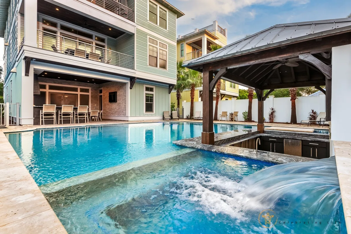 A vibrant exterior view features a private heated pool, a swim-up bar under a tiki-style pavilion, outdoor seating, and the modern facade of a multi-story beach house. The scene is lushly landscaped and bathed in sunlight, creating a lively, welcoming environment that invites outdoor relaxation, group activities, and pet-friendly enjoyment.