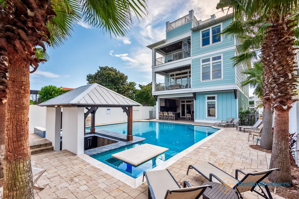 A spacious private heated pool glistens in the sun, complete with an inviting swim-up bar and adjacent poolside tiki kitchen. The pool area is framed by palm trees and features lounge chairs around the edges. The modern multi-story beach house with balconies forms the backdrop, tying in the luxury, privacy, and dog-friendly fencing mentioned in the narrative.