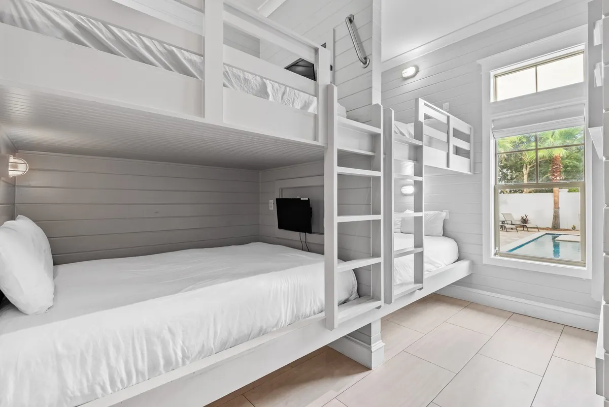A cheerful bunk room featuring built-in bunk beds with ladders, soft white bedding, and pillows. A window provides a view of the pool area, and wall-mounted reading lights offer personal comfort. The space is clearly designed for children or groups, offering both fun and privacy.