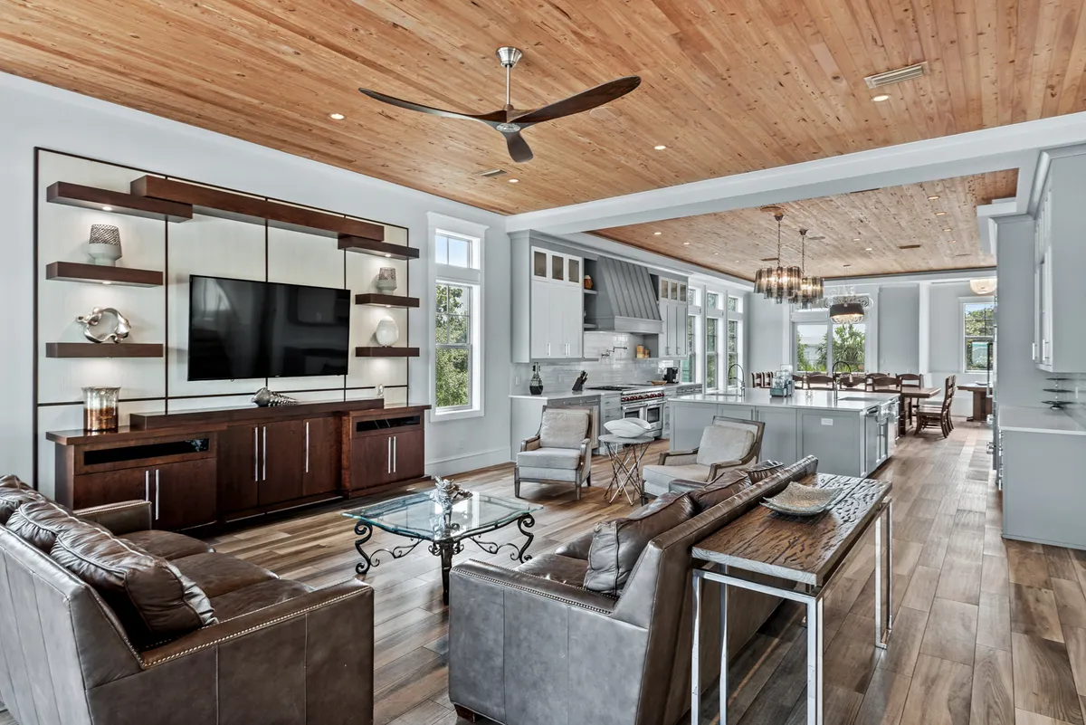 A spacious living area arranged for group relaxation, with a large flat-screen TV on custom wooden shelving, leather sofas around a glass coffee table, an open-concept kitchen with a large island and bar stools, and a dining area visible in the background. The setting is upscale, warm, and perfect for movie nights or casual storytelling after adventures.