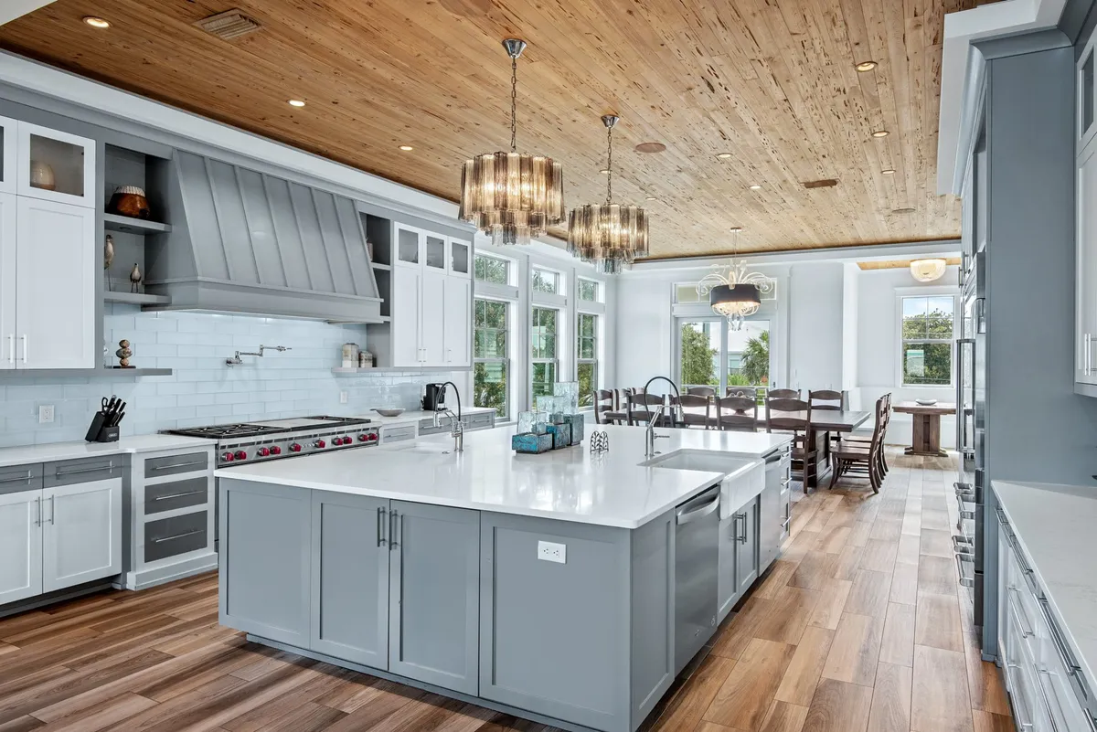This image showcases a spacious, open-concept kitchen and dining area. The scene features a large central kitchen island with a white countertop, modern stainless steel appliances—including a wide range stove—wood-paneled ceiling with recessed lighting and elegant chandeliers, and a dining area with a large wooden table and chairs positioned near windows that let in abundant natural light.
