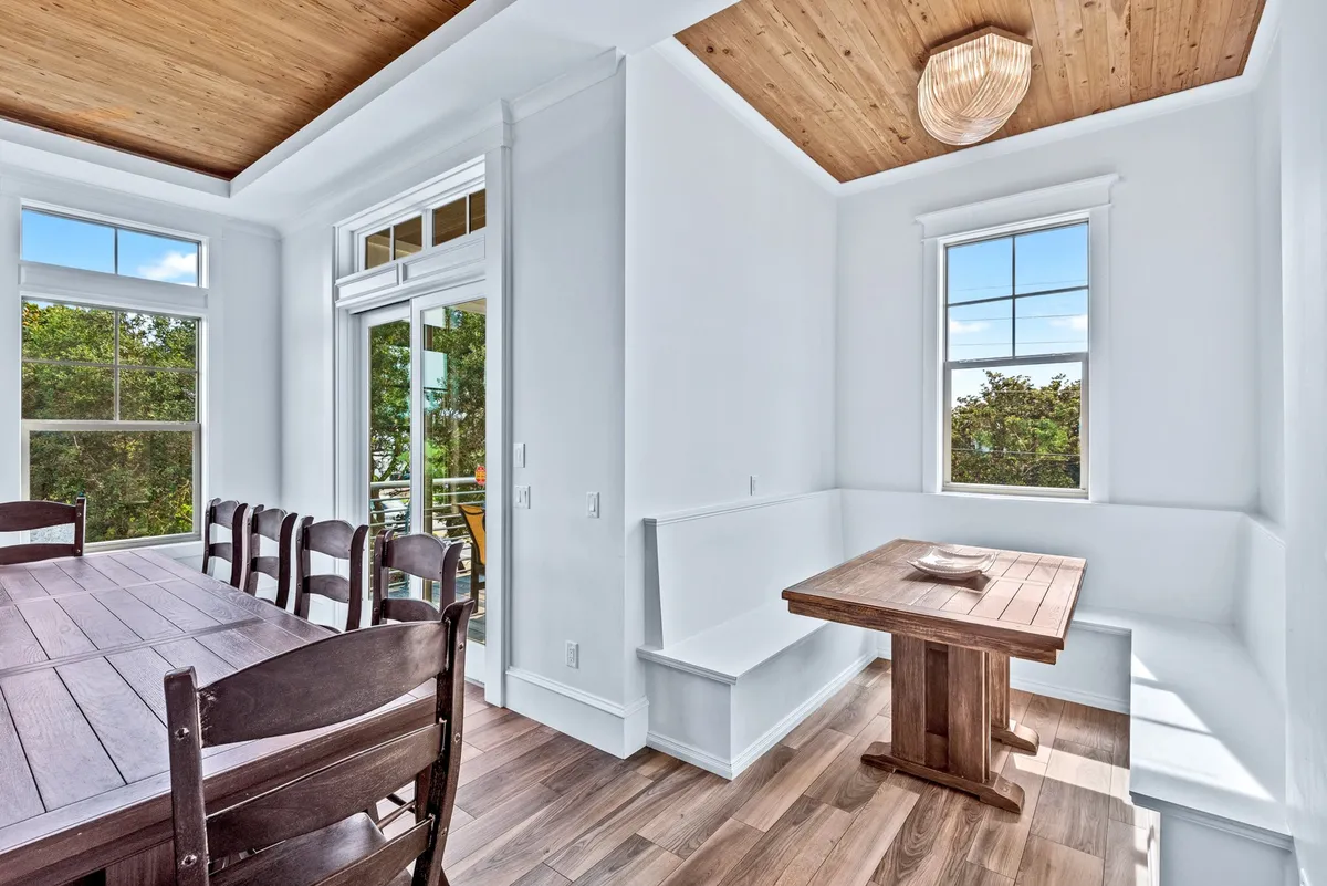 A cozy breakfast nook features built-in white corner bench seating around a wooden table, with additional chairs, large windows offering greenery views, and a wood-paneled ceiling with a unique light fixture. The mood is relaxed and intimate, ideal for casual breakfasts or late-night snacks.