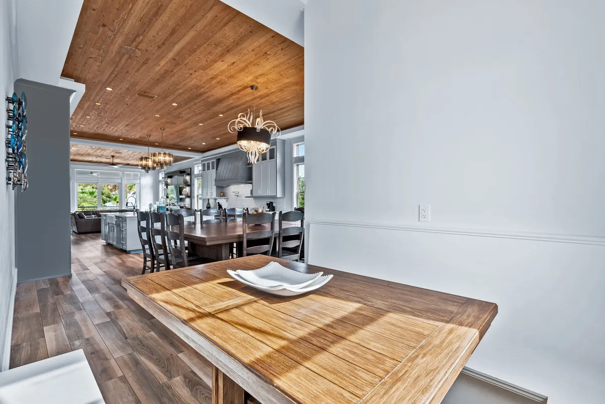 A warm, stylish dining area with a large wooden table surrounded by chairs, a kitchen island with additional seating, unique chandeliers, and an open kitchen equipped with white cabinetry and stainless steel appliances. The area is brightly lit by natural light, creating an inviting atmosphere for both large-group dinners and casual meals.