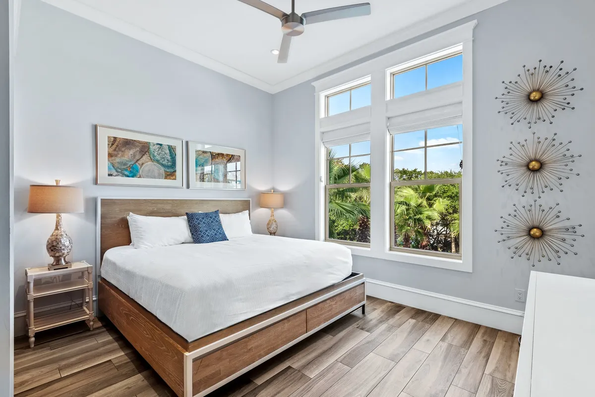 A large king-sized bed with pristine white linens anchors the room. Three decorative metal wall sculptures add a touch of elegance above the bed, while two bedside lamps with textured bases provide soft, ambient light. Expansive windows reveal lush tropical greenery outside, creating a serene and restful retreat.