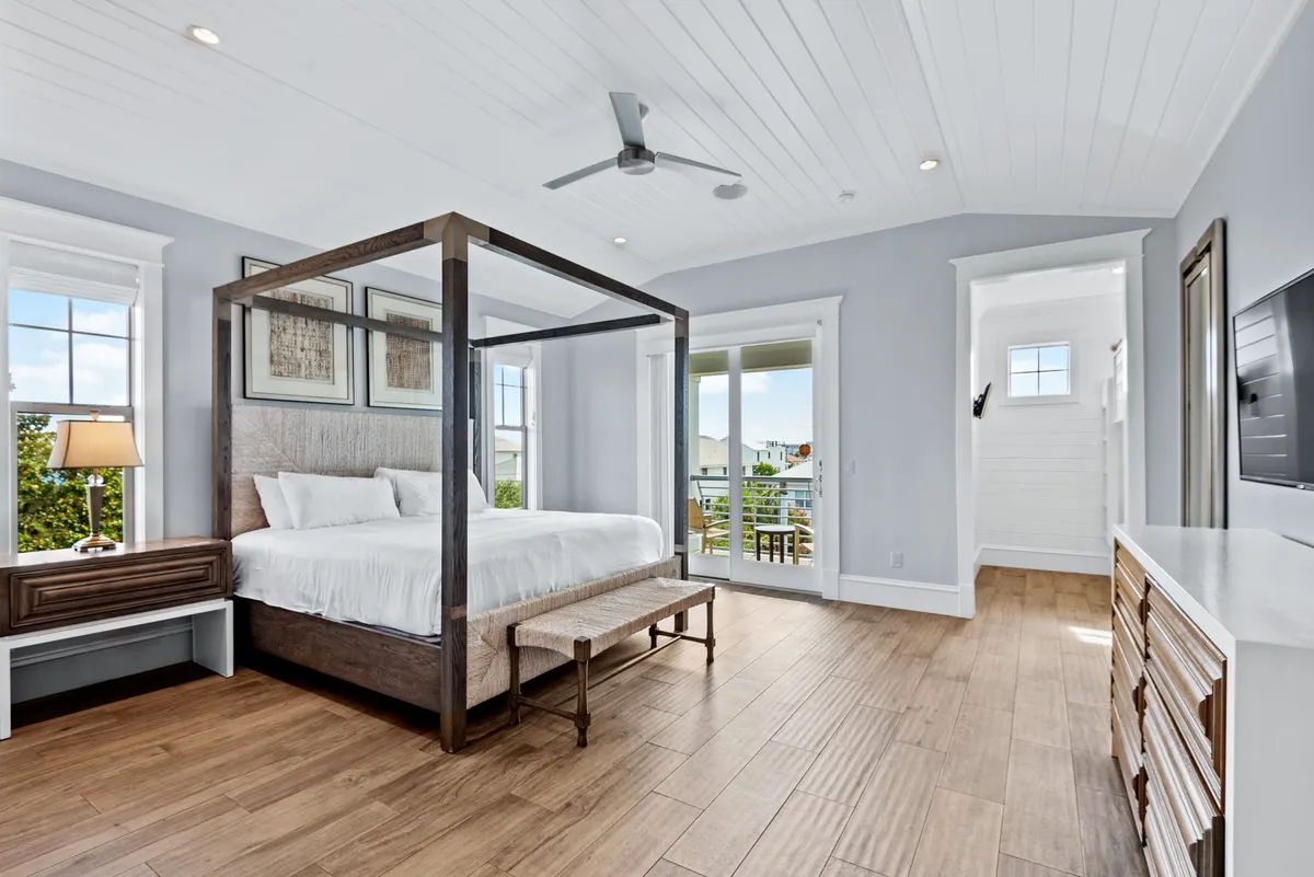 A spacious king bedroom with a four-poster bed and neutral bedding. Glass doors open to a private balcony with a poolside view, while natural wood flooring and a large dresser provide comfort and storage. Mounted flat screen TV and tranquil decor complete the retreat.