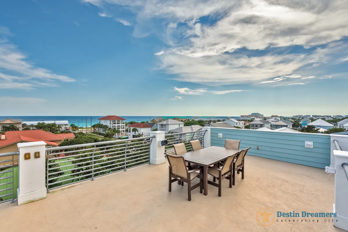 A rooftop patio with a dining table and chairs offers expansive Gulf and neighborhood views. Surrounded by a modern metal railing and blue siding wall, this serene space is ideal for sunset cocktails, morning coffee, or stargazing—exactly as described in the section's rooftop terrace feature.