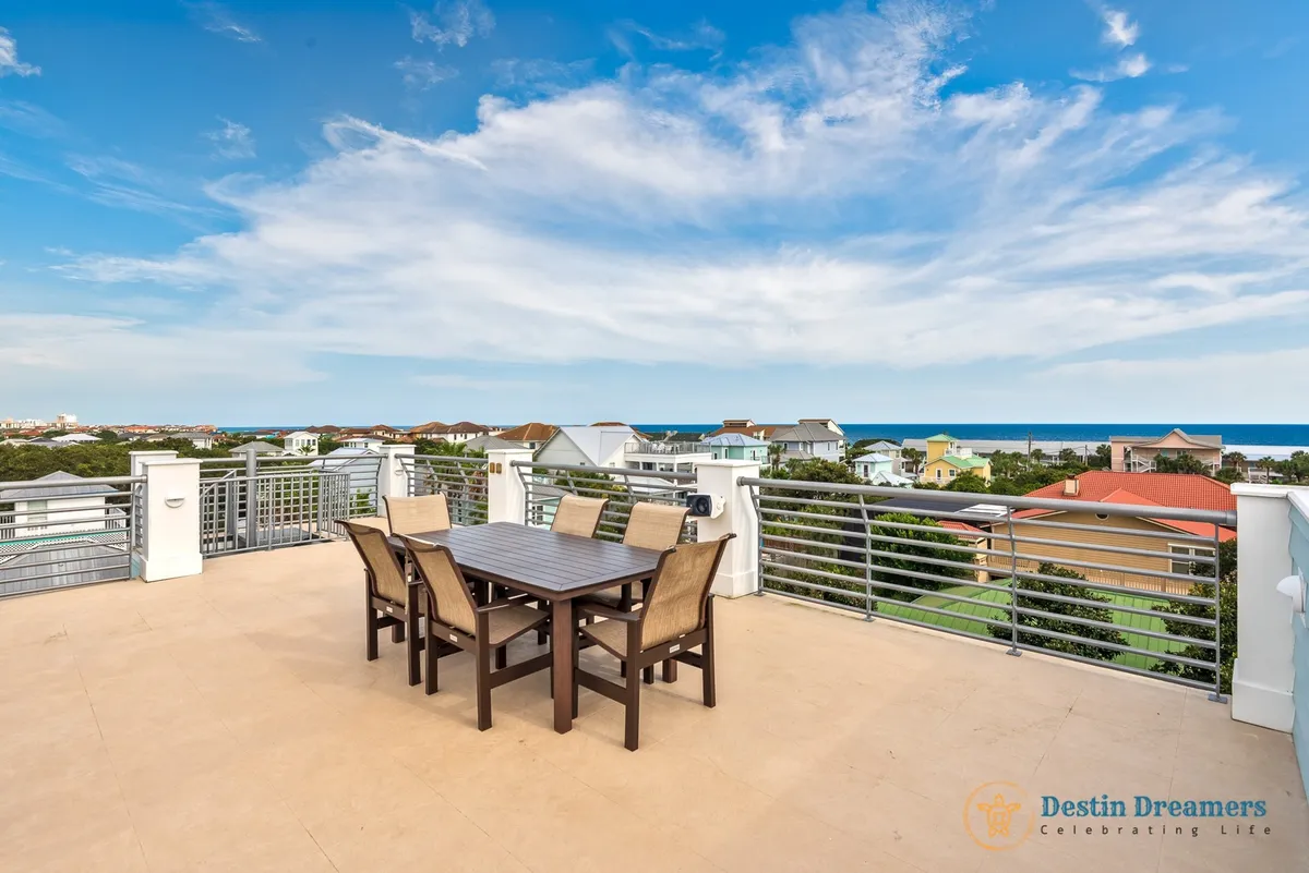 A rooftop patio set for al fresco dining, with a metal railing, six-seat outdoor dining table, and sweeping views of colorful rooftops and the distant Gulf of Mexico. The setting is tranquil and inviting, perfect for sunset gatherings or relaxed team socials.