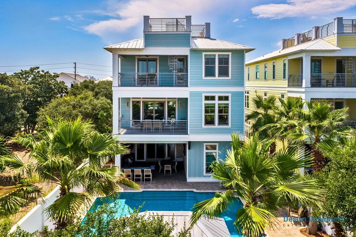 An exterior view of the property reveals a private heated pool with a swim-up bar, surrounded by lounge chairs and lush palm trees. The multi-story beach house features balconies and a rooftop patio, hinting at luxurious amenities and space for family gatherings. Outdoor dining and seating spaces are visible, reinforcing the blend of relaxation and social connection.