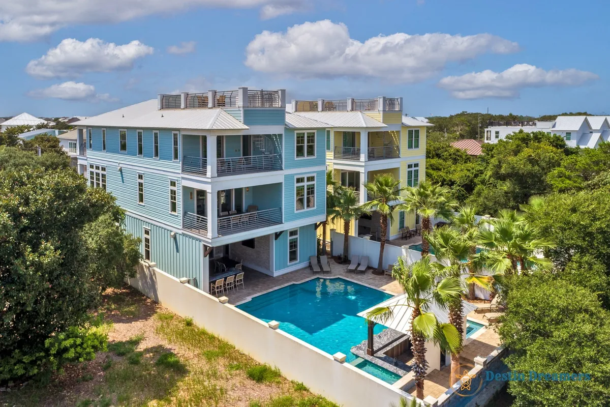 A vibrant exterior shot of The Life Aquatic, showcasing the three-story coastal-style house with multiple balconies, a private sunken swim-up bar and cabana beside the pool, lush palm trees, and ample outdoor lounge seating. The scene is lively yet tranquil, capturing the property's upscale amenities and its inviting group spaces.