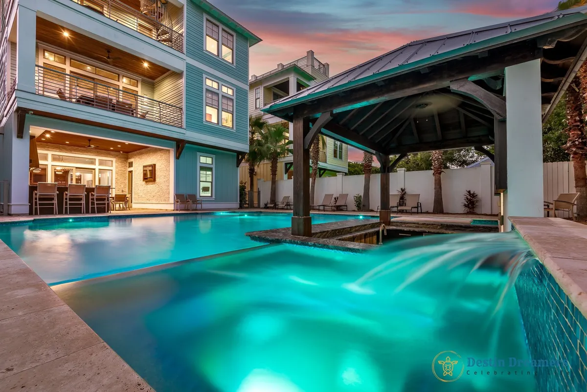 A captivating evening scene: the private heated pool with a sunken swim-up bar, covered tiki bar structure, and multi-story beach house in the background. Ambient pool lighting creates a relaxing glow, while outdoor seating and dining areas showcase the perfect setup for group celebrations and sunset cocktails.