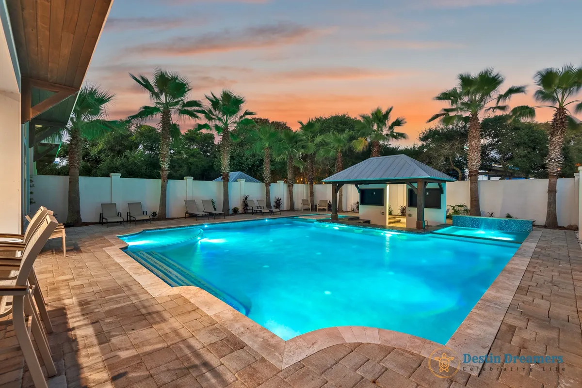 A luxurious heated private pool with a sunken swim-up bar is the centerpiece, surrounded by a stone-tiled pool deck, palm trees, and a tiki bar pavilion. Lounge chairs line the poolside, creating an inviting outdoor oasis. The design and openness suggest a space perfect for both family relaxation and group gatherings, with ample room for pets to enjoy the outdoors as well.